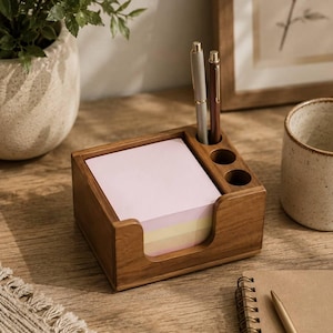 May include: A rectangular wooden desk organiser featuring a square compartment filled with a stack of light pink and yellow sticky notes, alongside three cylindrical holes designed to hold pens, with two pens currently inserted in the holder.