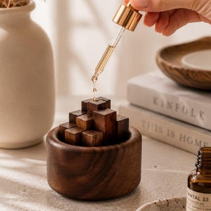 May include: A small, round, dark brown wooden bowl filled with a cluster of stacked wooden cubes. A glass dropper with a gold-colored cap is positioned above the cubes, releasing a single drop of clear liquid onto the center of the wooden structure.