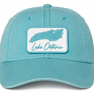 Op de afbeelding: Een turquoise honkbalpet met een rechthoekige witte patch aan de voorzijde. De patch toont een geborduurde turquoise omtrek van het Lake Ontario boven de tekst Lake Ontario in een cursief turquoise lettertype met een bijpassende rand.
