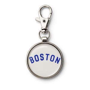 May include: A circular silver-toned metal keychain charm featuring a white insert with the word BOSTON printed in blue capital letters in an arched font. The charm is attached to a silver-toned metal lobster claw clasp for easy attachment.