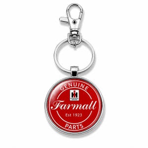 May include: A round silver keychain featuring a red circular emblem with white text that reads Genuine Farmall Parts Est 1923 and the International Harvester logo in the center. The keychain includes a metal split ring and a lobster claw clasp.