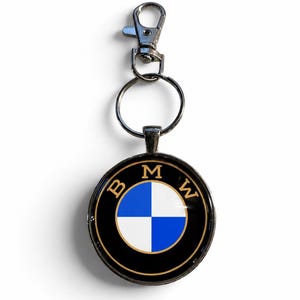 May include: A circular keychain featuring the BMW logo with a black outer ring, gold lettering, and a blue and white checkered center. The pendant is attached to a silver-toned metal split ring and a lobster claw clasp for secure attachment.