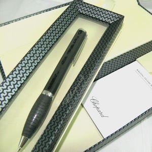 May include: A black Chopard ballpoint pen with a silver clip and a textured grip section, presented in a rectangular box with a black and white wavy pattern. A white card with the Chopard logo and the text WRITING INSTRUMENT is included.