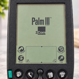 May include: A black 3Com Palm III personal digital assistant featuring a monochrome screen displaying the Palm III and 3Com logos. The device includes a navigation rocker, four circular application buttons, a green power button, and a stylus input area.