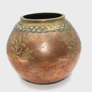 May include: A round copper vase featuring a wide brass rim with a decorative scalloped edge pattern. The body of the vase is etched with delicate floral designs, showcasing a warm metallic finish with visible signs of natural patina and wear.