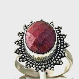 May include: A silver ring featuring a large, oval-shaped, faceted deep red gemstone set in a bezel. The setting is surrounded by a decorative sunburst-style border composed of small, raised silver beads, creating a textured, metallic frame.