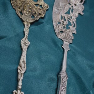 Puede incluir: Un tenedor y un cuchillo de servir de metal en tono plateado. El tenedor presenta un mango ornamentado y una cabeza de tres puntas con un medallón central. El cuchillo tiene una hoja ancha y curva con intrincados calados y motivos florales.