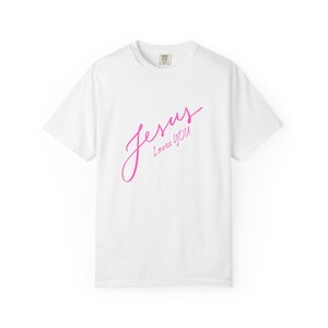 May include: A white short-sleeved t-shirt featuring the text Jesus Loves You printed in a bright pink cursive script across the center of the chest. The garment has a crew neckline and a standard fit, showcasing a clean and simple design.