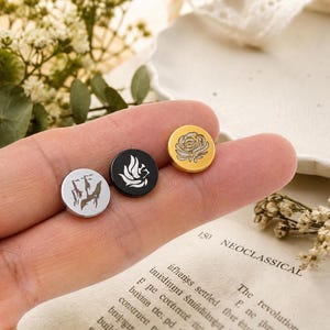 Puede incluir: Tres pines metálicos circulares con grabados distintos: un pin plateado con un paisaje de montaña, uno negro con un diseño de ala estilizada y uno dorado con una ilustración detallada de una rosa.