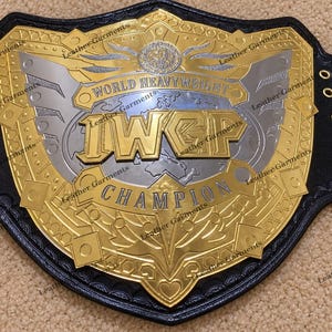 Può includere: Una cintura da campionato con una grande placca centrale dorata e argentata con la scritta WORLD HEAVYWEIGHT IWGP CHAMPION. La placca è montata su un cinturino in pelle nera con occhielli metallici dorati sui lati e incisioni decorative.