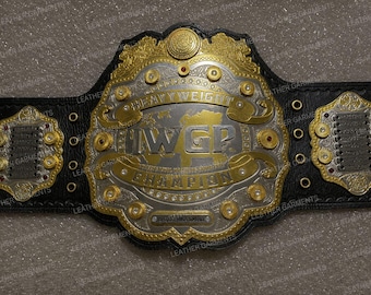 Pode incluir: Um cinturão de campeonato de couro preto com uma grande placa central dourada e prateada com o texto IWGP HEAVYWEIGHT CHAMPION e um design de mapa-múndi. O cinto inclui duas placas laterais menores com detalhes prateados e dourados.