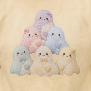 May include: A pale yellow t-shirt featuring a central graphic of five stacked, pastel-coloured ghost figures. Each ghost has a smiling face, a small bow, and a heart-shaped patch, rendered in shades of lavender, pink, blue, cream, and yellow.