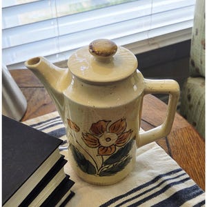 May include: A cream-colored ceramic teapot featuring a hand-painted orange flower with dark green leaves on the front. The teapot has a matching lid with a rounded knob, a sturdy handle, and a spout. The surface has a subtle crackle glaze finish.
