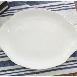 May include: A round, white ceramic plate featuring a scalloped edge with subtle embossed detailing along the rim. The surface of the plate is smooth and glossy, with a clean, solid white finish that highlights the gentle curves of the border.