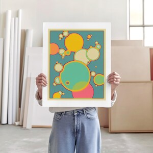 A person is holding up a framed artwork with a colorful abstract design featuring circles and shapes in various shades of green, yellow, and pink.