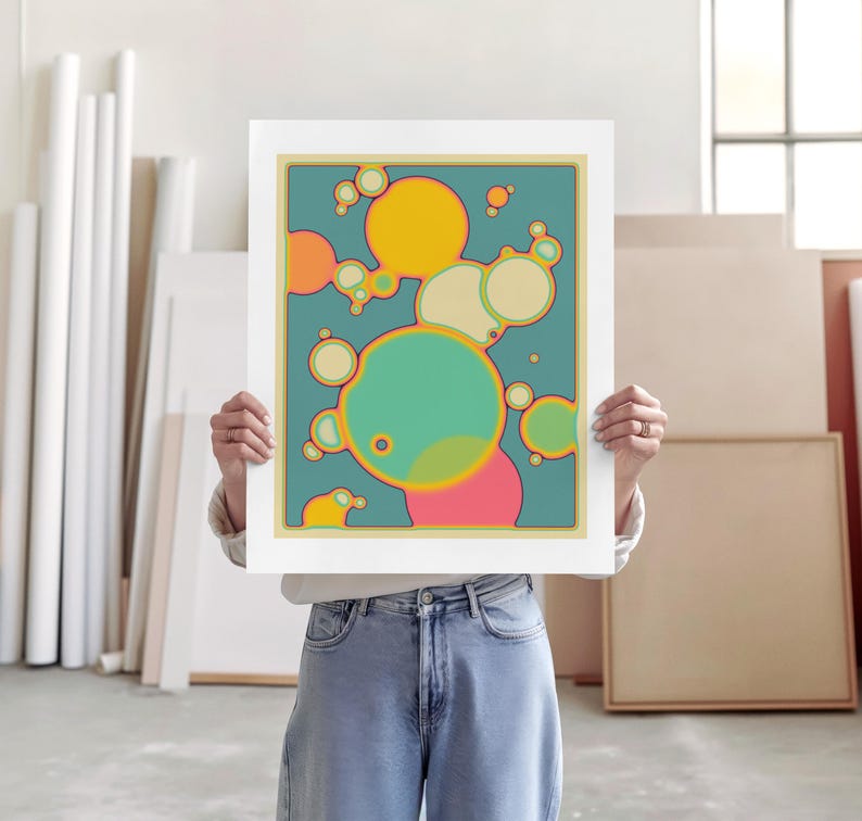 A person is holding up a framed artwork with a colorful abstract design featuring circles and shapes in various shades of green, yellow, and pink.