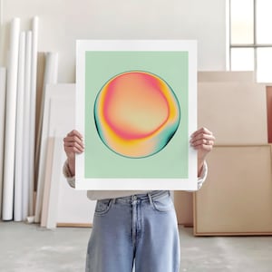 A person is holding up a large square poster with a colorful abstract design on it, standing in a room with various art supplies and materials visible in the background.