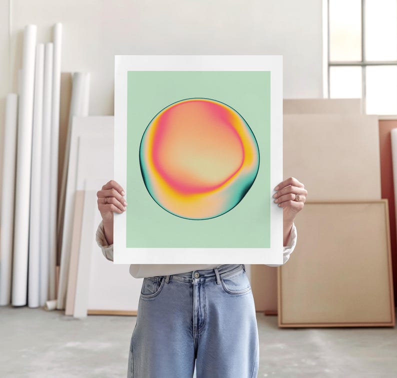 A person is holding up a large square poster with a colorful abstract design on it, standing in a room with various art supplies and materials visible in the background.