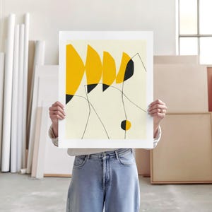 A person is holding up a white piece of paper with a yellow abstract design on it, standing in a room with various art supplies and materials visible in the background.