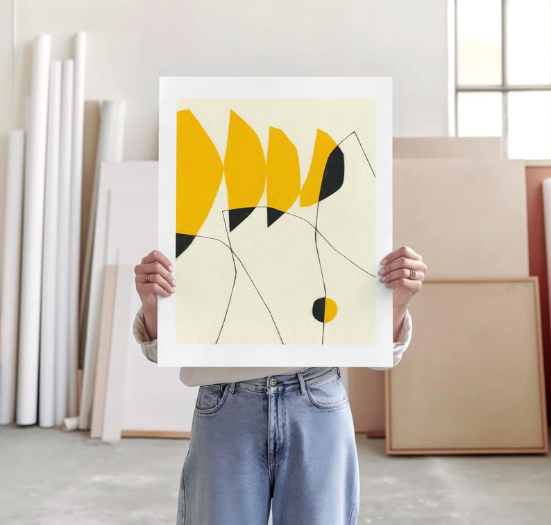 A person is holding up a white piece of paper with a yellow abstract design on it, standing in a room with various art supplies and materials visible in the background.