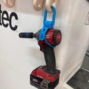 May include: A red and black Milwaukee M18 Fuel impact driver with a black battery pack, featuring a One-Key label and a black socket attached to the front. The tool is suspended from a white hook by a bright blue plastic hanger accessory.