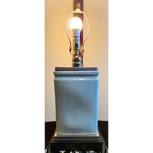 May include: A table lamp featuring a rectangular, light blue ceramic base with a smooth, glossy finish. The lamp has a metallic silver socket assembly holding a single glowing light bulb, mounted on a dark, square base stand.