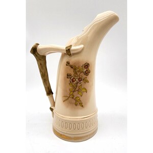 May include: A cream-colored ceramic pitcher with a horn-shaped body and a handle textured to resemble a branch. The front features a painted floral design with green leaves and brown-centered flowers, and the base has a molded ring pattern.
