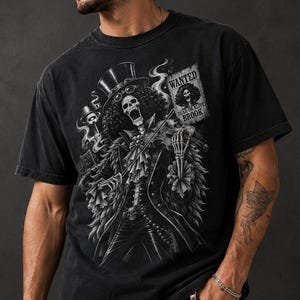 May include: A black short-sleeve t-shirt featuring a graphic print of a skeleton character wearing a top hat and playing a violin. The design includes a 'WANTED DEAD OR ALIVE BROOK' poster. The shirt is labeled Comfort Colors 1717 in black.