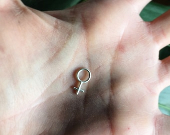 May include: A silver charm in the shape of the female gender symbol. The charm is small and delicate, and is shown resting in the palm of a hand.