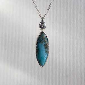 May include: A silver chain necklace with a pendant featuring a blue and gray gemstone set in a silver bezel. The pendant has a small diamond accent at the top.