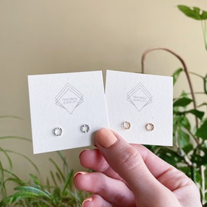 May include: Two cards with the text "Tiny Erica Jewelry" on them. Each card has two small earrings on them. The earrings on the left card are silver and the earrings on the right card are gold.