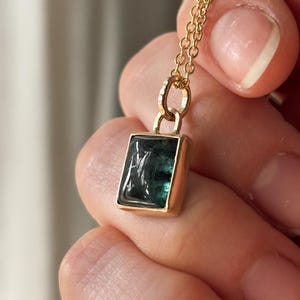 May include: A gold necklace with a rectangular, dark green gemstone pendant. The pendant is set in a gold frame and hangs from a delicate gold chain. The gemstone has a slightly translucent appearance.