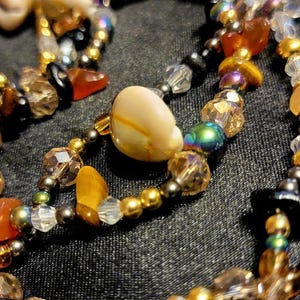 May include: A multi-strand beaded necklace featuring a variety of materials including white cowrie shells, faceted clear crystals, metallic gold beads, dark grey metallic beads, and irregular orange and brown stone chips arranged in a layered design.