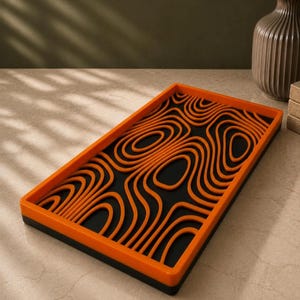May include: A rectangular tray featuring a black base with raised orange lines in a topographic map pattern. The tray has a solid orange outer rim and a black underside, presenting a high-contrast design with a layered, three-dimensional appearance.