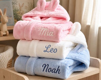 May include: A stack of three plush hooded robes in pink, white, and light blue. Each robe features a hood with two small ears and is embroidered with a name: Mia on the pink robe, Leo on the white robe, and Noah on the light blue robe.