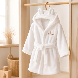 May include: A white plush bathrobe featuring a hood with two small rounded ears on top. The robe has long sleeves, a matching tie belt at the waist, and the name Ava embroidered in gold cursive script on the left side of the chest area.
