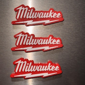 Può includere: Tre emblemi identici in plastica rossa e bianca con il logo del marchio Milwaukee. Ogni pezzo mostra la parola Milwaukee in un carattere corsivo bianco stilizzato su sfondo rosso, con una grafica a forma di fulmine bianco sottostante.
