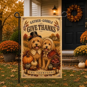 May include: A rectangular garden flag featuring two golden-coloured dogs dressed in pilgrim-style clothing. The text reads GATHER GOBBLE GIVE THANKS and THE THOMPSONS. The illustration includes pumpkins, gourds, and a basket of apples on a field background.