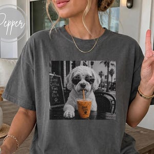 May include: A charcoal gray crewneck t-shirt featuring a black and white graphic print of a dog wearing sunglasses and drinking iced coffee. The graphic includes a sign that reads Iced Coffee Fuel the Pack and a cup labeled Good Vibes.