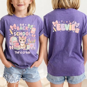Puede incluir: Una camiseta morada con un gráfico frontal de un gato con gafas, material escolar y el texto Back to School Third Grade. La parte trasera de la camiseta muestra el nombre Eevie en letras grandes rodeado de lazos, estrellas y un lápiz.