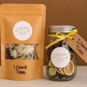 Könnte beinhalten: Ein brauner Papierbeutel und ein Glasgefäß gefüllt mit getrockneten Früchten und botanischen Zutaten. Beide haben ein weißes, rundes Etikett mit dem Text simply, katie made simply loved deeply und einen braunen Anhänger mit dem Text Island Time.