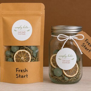 May include: A brown kraft paper pouch and a clear glass mason jar, both filled with dried eucalyptus leaves and orange slices. Each container features a white circular label reading simply, katie made simply, loved deeply and the text Fresh Start.