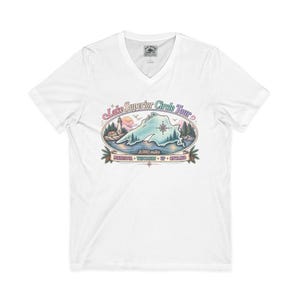Peut inclure: Un t-shirt blanc à col en V avec un motif représentant le lac Supérieur, un phare, une boussole et des pins. Le texte indique Lake Superior Circle Tour, 1300 miles, Minnesota, Wisconsin, UP, Ontario. Le design utilise des tons pastel doux.