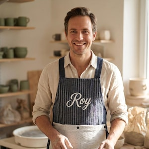 May include: A dark blue pinstriped apron featuring the name Roy embroidered in white script across the chest, with a white textured pocket panel at the bottom secured by two dark buttons, worn over a light beige button-down shirt.
