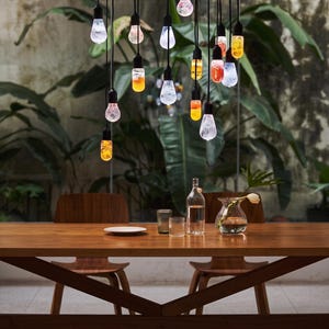 May include: A multi-light pendant chandelier featuring numerous hanging bulbs with textured glass in shades of orange, red, and clear white, suspended by black cords above a wooden dining table with two chairs, a glass bottle, and a vase.