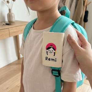 Peut inclure: Une étiquette rectangulaire en tissu couleur crème fixée sur la bretelle d'un sac à dos bleu sarcelle vif. L'étiquette présente une illustration brodée d'une personne portant un chapeau rose et le prénom Remi cousu en fil noir.