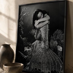 May include: A black and white framed wall art print depicting a mermaid with long dark hair, sitting on a rock formation. The mermaid is shown with arms crossed, resting on a textured tail covered in scales, set against a dark underwater background.