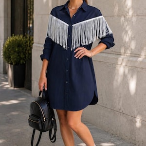 May include: A navy blue button-down shirt dress featuring long sleeves, a classic collar, and white fringe detailing across the chest and shoulders. The dress has a curved hemline and is paired with white athletic trainers, white socks, and a black backpack.