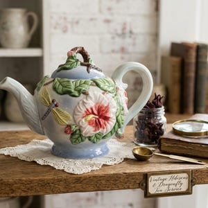 May include: A ceramic teapot with a light blue glaze, featuring three-dimensional sculpted hibiscus flowers, green leaves, and a dragonfly. The lid has a branch-like handle. A brass label on the shelf reads Vintage Hibiscus & Dragonfly Tea Service.