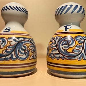 May include: A pair of ceramic salt and pepper shakers with a rounded, bulbous shape. Each shaker features a white base with hand-painted blue and yellow scrollwork patterns. The letter S is marked on one shaker, and the letter P is on the other.